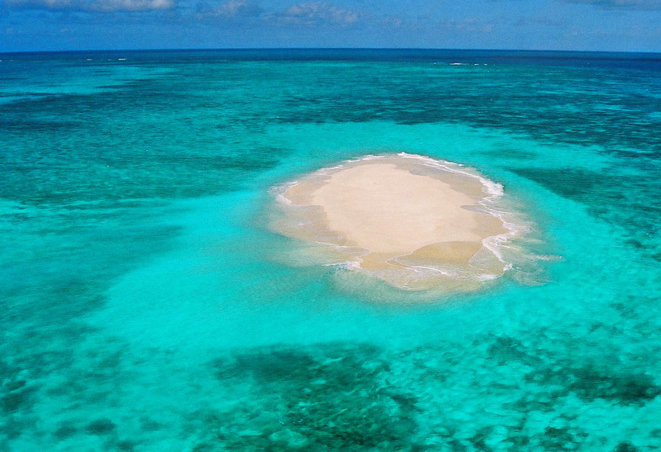 A small sandy island surrounded by clear turquoise ocean water under a blue sky.
