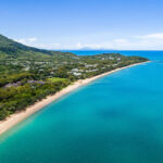 Aerial view of a coastline with turquoise water, sandy beach, lush green trees, and buildings nestled near a forested hill under a clear blue sky.