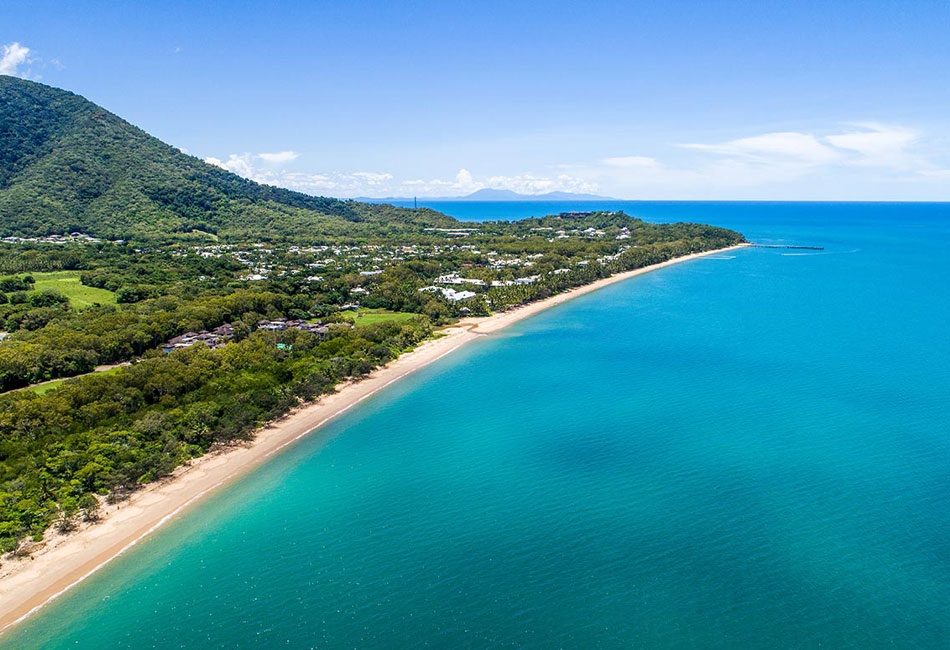Aerial view of a coastline with turquoise water, sandy beach, lush green trees, and buildings nestled near a forested hill under a clear blue sky.