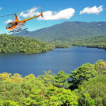 An orange helicopter flies above a lush, green forest and a large blue lake with mountains in the background under a clear sky.