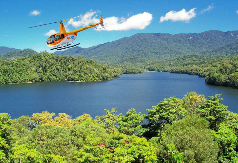 An orange helicopter flies above a lush, green forest and a large blue lake with mountains in the background under a clear sky.