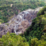 A narrow waterfall flows down rocky terrain surrounded by dense green forest, with the water appearing to trickle in several sections.