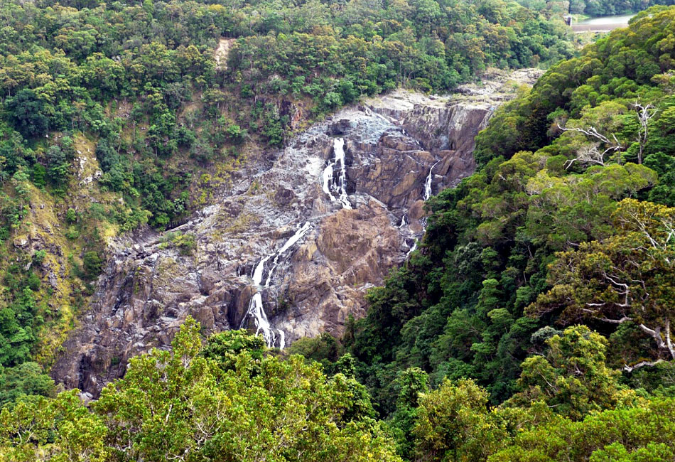 A narrow waterfall flows down rocky terrain surrounded by dense green forest, with the water appearing to trickle in several sections.