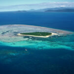 A small, green island surrounded by sandy beaches, shallow turquoise water, and coral reefs, with deeper blue ocean and distant islands in the background.