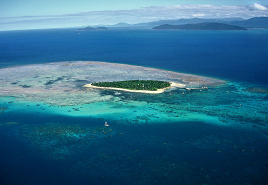 A small, green island surrounded by sandy beaches, shallow turquoise water, and coral reefs, with deeper blue ocean and distant islands in the background.