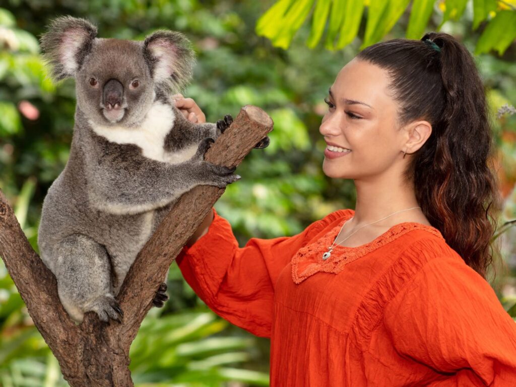 A woman in an orange blouse stands next to a koala perched on a tree branch in a lush, green outdoor setting.