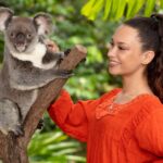 A woman in an orange blouse stands next to a koala perched on a tree branch in a lush, green outdoor setting.