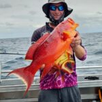 A person wearing a hat and sunglasses holds a large, bright orange fish on a boat at sea under a cloudy sky.