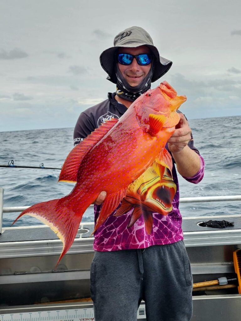 A person wearing a hat and sunglasses holds a large, bright orange fish on a boat at sea under a cloudy sky.
