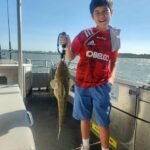 A boy on a boat holds up a large fish by the mouth with a grip tool, smiling at the camera.