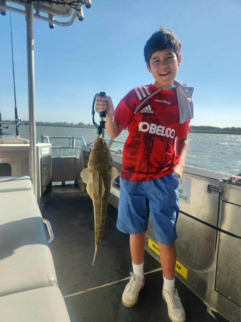 A boy on a boat holds up a large fish by the mouth with a grip tool, smiling at the camera.