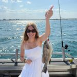 A woman in a white dress and sunglasses holds up a fish she caught on a fishing boat, with water and distant buildings in the background.