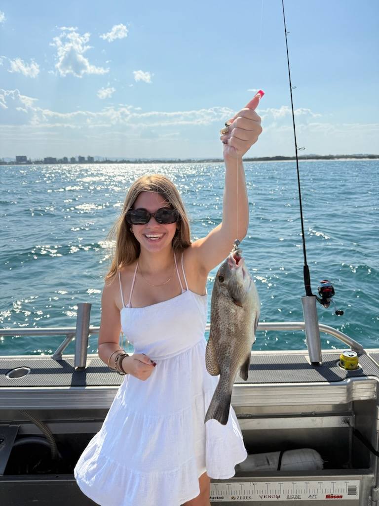 A woman in a white dress and sunglasses holds up a fish she caught on a fishing boat, with water and distant buildings in the background.