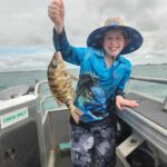 A smiling child on a boat holds up a freshly caught fish. The child is wearing a blue fishing shirt, hat, and shorts. Water and cloudy sky are visible in the background.