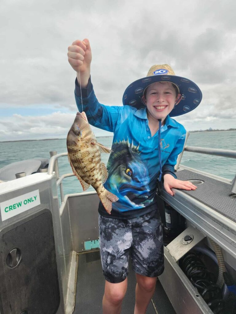 A smiling child on a boat holds up a freshly caught fish. The child is wearing a blue fishing shirt, hat, and shorts. Water and cloudy sky are visible in the background.