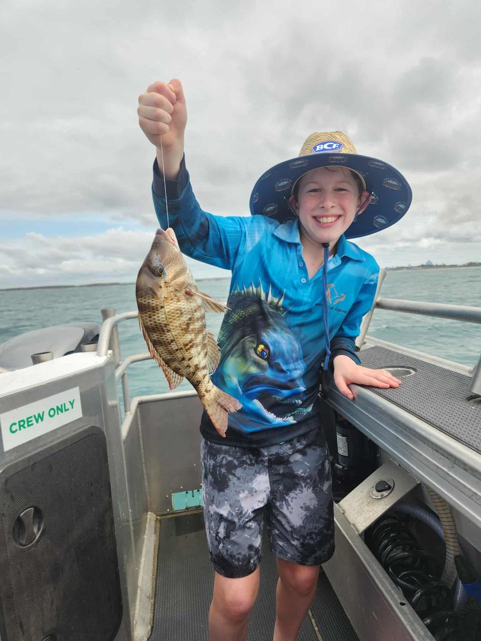 A smiling child on a boat holds up a freshly caught fish. The child is wearing a blue fishing shirt, hat, and shorts. Water and cloudy sky are visible in the background.
