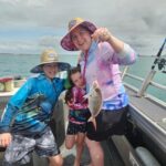 Three people on a boat; the adult holds a fish on a line. They are all smiling and wearing hats and sun-protective clothing. The sea and cloudy sky are in the background.