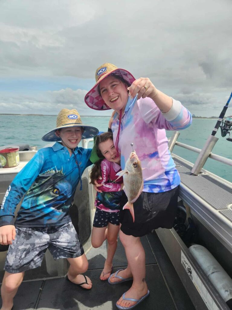 Three people on a boat; the adult holds a fish on a line. They are all smiling and wearing hats and sun-protective clothing. The sea and cloudy sky are in the background.