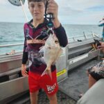 A boy standing on a boat holds up a fish with a clamp tool. Fishing net and ocean are visible in the background.