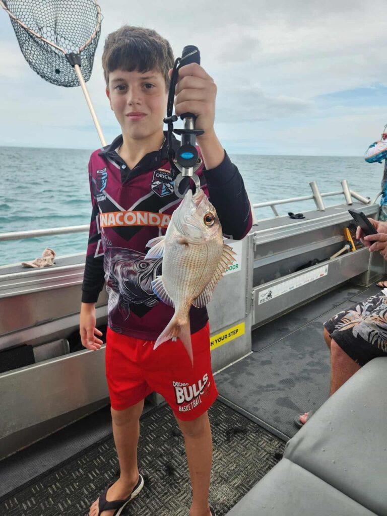 A boy standing on a boat holds up a fish with a clamp tool. Fishing net and ocean are visible in the background.