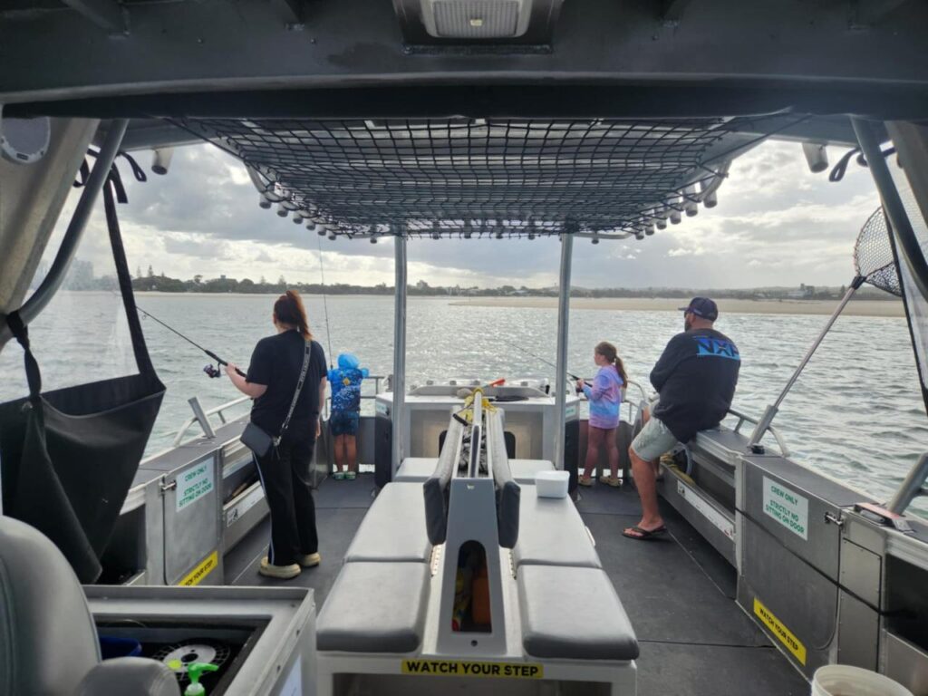Four people, including two children, are fishing on a boat in calm water under a cloudy sky. Safety signs and gear are visible on the boat.