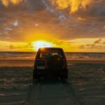 A red SUV is parked on a sandy beach facing the ocean, with tire tracks behind it and a dramatic sunset sky overhead.