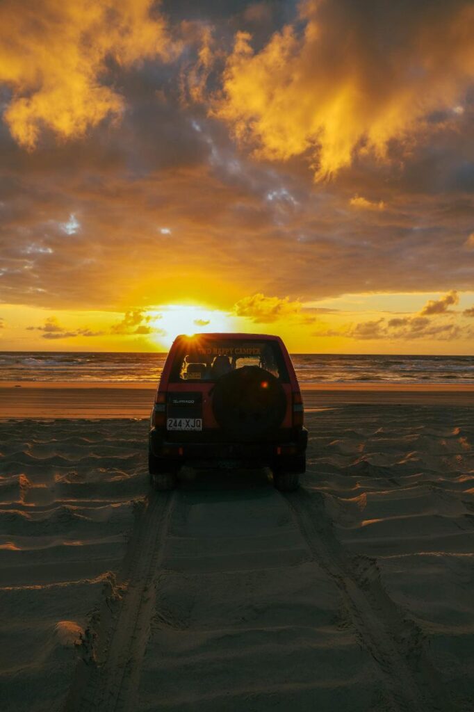 A red SUV is parked on a sandy beach facing the ocean, with tire tracks behind it and a dramatic sunset sky overhead.
