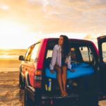 A woman sits in the back of an SUV parked on a sandy beach at sunset, with a blue mattress and sunlight reflecting on the water.