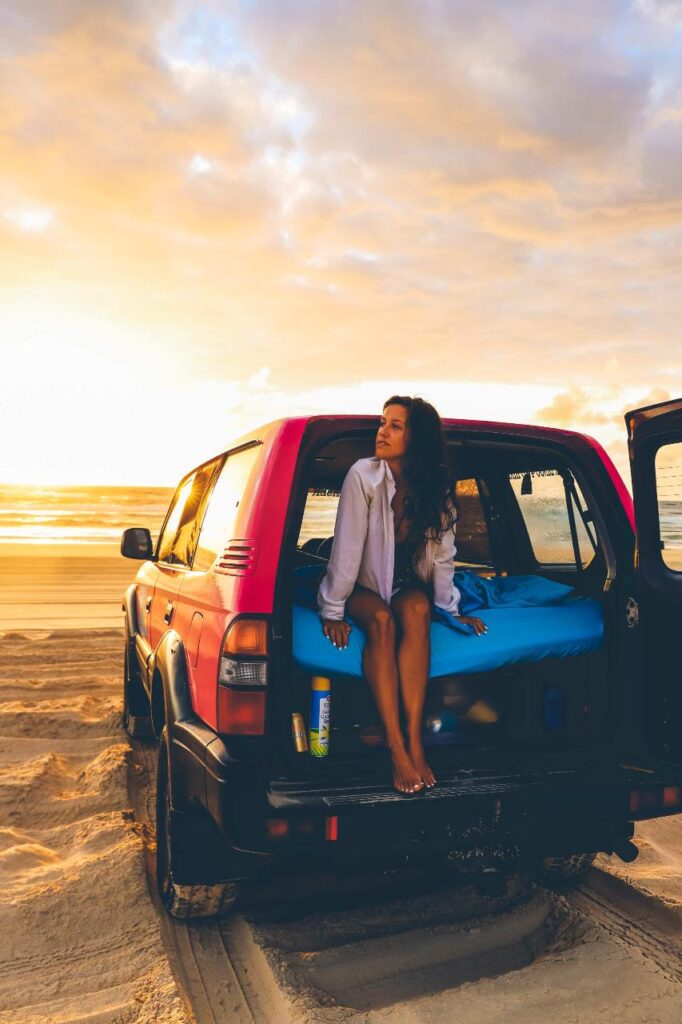 A woman sits in the back of an SUV parked on a sandy beach at sunset, with a blue mattress and sunlight reflecting on the water.