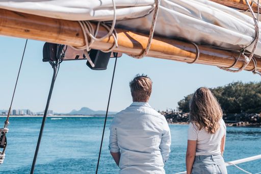 Two people, seen from behind, stand on a boat looking out over calm blue water with land and trees in the distance.