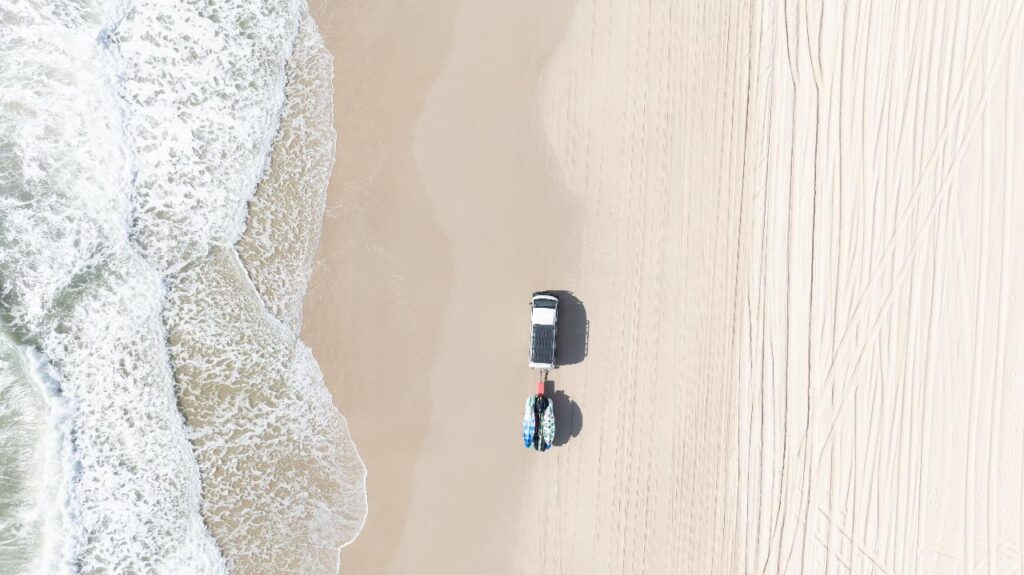 Aerial view of a white truck parked on a sandy beach near the shoreline, with surfboards attached to its rear and tire tracks visible in the sand.
