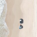 Aerial view of a white truck parked on a sandy beach near the shoreline, with surfboards attached to its rear and tire tracks visible in the sand.