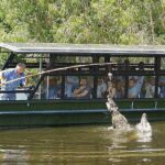 A man on a tour boat uses a stick to interact with a jumping crocodile in the water, as passengers watch from behind protective bars.
