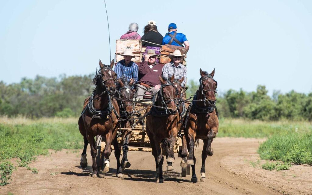 A group of people rides in a wooden wagon pulled by four horses down a dirt road, surrounded by greenery under a clear sky.