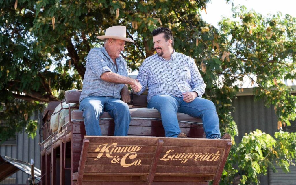 Two men in casual shirts and hats shake hands while sitting on a wooden wagon with “Kinnon & Co. Longreach” written on the back, with trees and a building in the background.