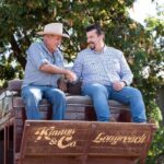 Two men in casual shirts and hats shake hands while sitting on a wooden wagon with “Kinnon & Co. Longreach” written on the back, with trees and a building in the background.