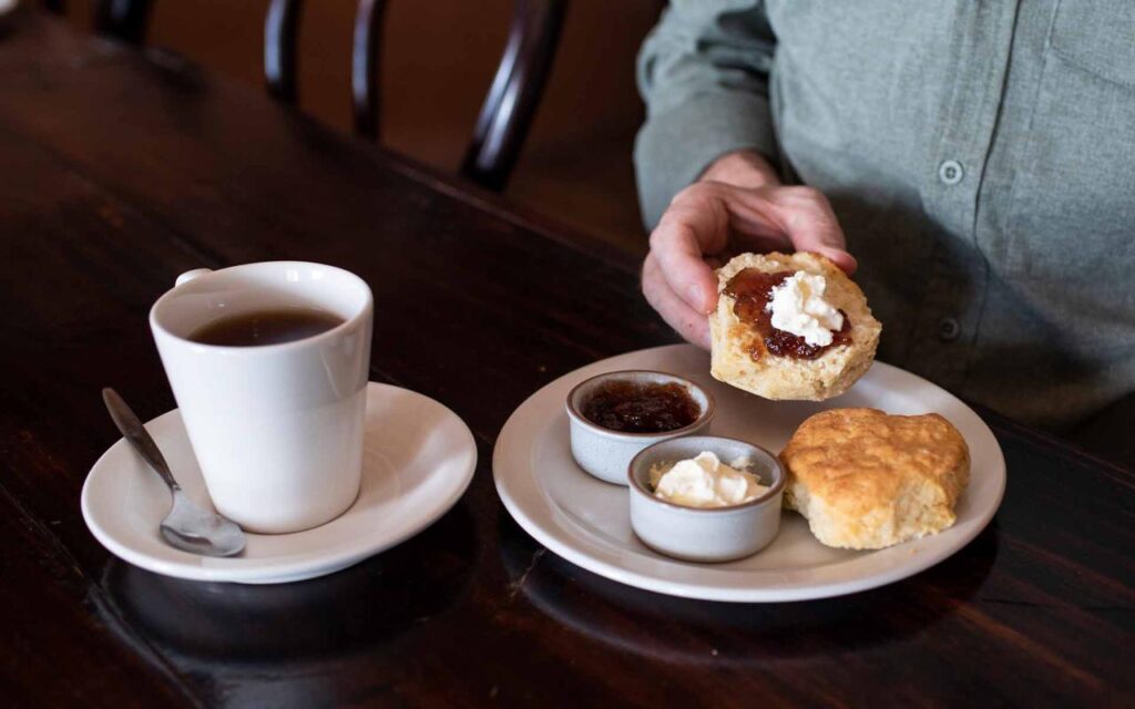 A person prepares a biscuit with jam and cream at a table, alongside a cup of tea, a spoon, and small bowls of jam and cream on a plate.