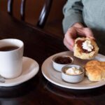 A person prepares a biscuit with jam and cream at a table, alongside a cup of tea, a spoon, and small bowls of jam and cream on a plate.