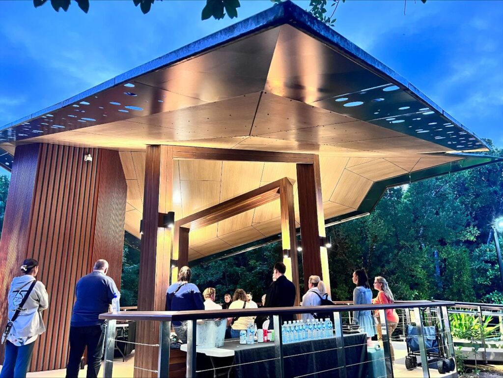 People gather under a modern, geometric pavilion at dusk. Bottled water and snacks are set on tables in the foreground; trees and foliage surround the scene.