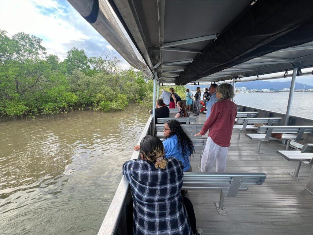 A group of people stands and sits on a boat, looking out at mangroves and water under a partly cloudy sky.