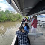 A group of people stands and sits on a boat, looking out at mangroves and water under a partly cloudy sky.