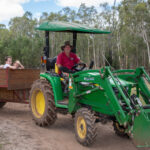 A man drives a green tractor pulling a wooden trailer filled with children, on a dirt path surrounded by trees.