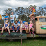 A group of children and two adults sit and stand on an old farm truck and bench outdoors, with trees and cloudy sky in the background.