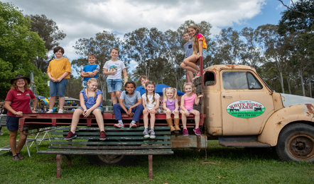 A group of children and two adults sit and stand on an old farm truck and bench outdoors, with trees and cloudy sky in the background.