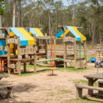 Outdoor playground with wooden play structures, picnic tables, toy cars, and a fenced area, surrounded by trees on a sunny day.