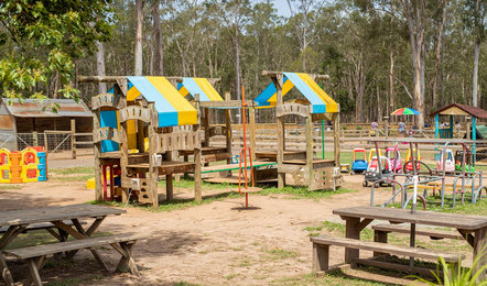 Outdoor playground with wooden play structures, picnic tables, toy cars, and a fenced area, surrounded by trees on a sunny day.