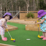 Two young children wearing hats and summer clothes play mini golf on a green outdoor course surrounded by wooden borders and trees.