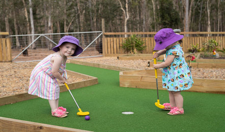 Two young children wearing hats and summer clothes play mini golf on a green outdoor course surrounded by wooden borders and trees.