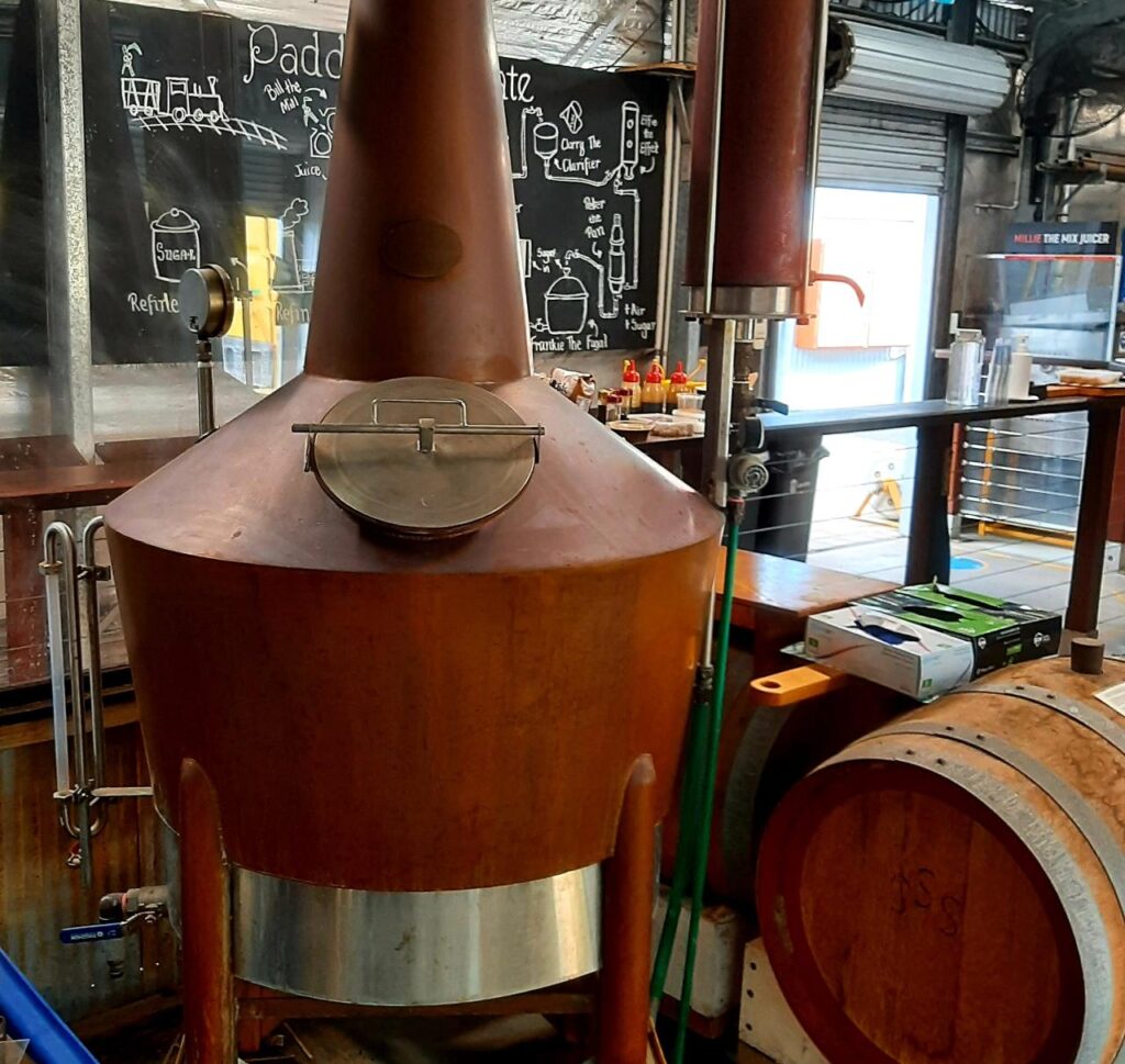 A copper brewing kettle with a closed lid stands next to a wooden barrel in a brewery; chalkboard instructions are visible in the background.