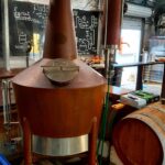 A copper brewing kettle with a closed lid stands next to a wooden barrel in a brewery; chalkboard instructions are visible in the background.